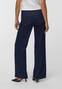 Široke kavbojke v temno indigo denim materialu, z višjim pasom, dvema zadnjima žepoma in diskretnim šivi. Skombinirane z rjavimi špici čevlji.