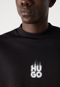 Fekete kapucnis pulóver, kerek nyakkal és texturált anyaggal, a mellkason kiemelkedő fehér "HUGO" logóval, lángszerű dizájnnal.