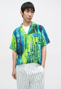 Koszula z krótkim rękawem z żywym, niebiesko-zielonym abstrakcyjnym wzorem, z zapięciem na guziki z przodu i lewej kieszeni. Noszona z białymi spodniami w paski.
