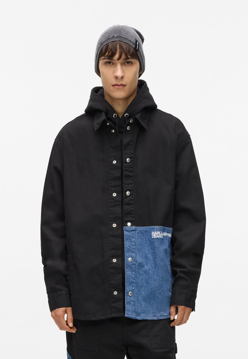 Melna kapuce jaka ar džinsu kabatu, ar pogu aizdari un "KARL LAGERFELD JEANS" logo. Tekstūra: gluda auduma un džinsa kontrasts.
