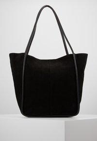 Svart mocka tote-väska med långa läderhandtag som vilar på en vit yta mot en enkel bakgrund.