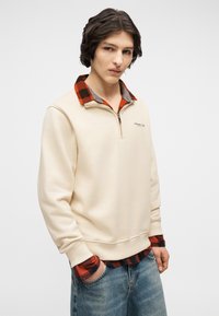 Νέος άνδρας στέκεται με τα χέρια στις τσέπες, φοράει εκρού μπλούζα πάνω από κόκκινο και μαύρο καρό πουκάμισο και μπλε τζιν, σε απλό φόντο.