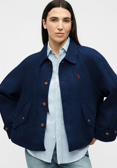 Γυναίκα που φορά χαλαρό σκούρο μπλε κουμπωτό σακάκι πάνω από ανοιχτό μπλε πουκάμισο και τζιν, στέκεται με τα χέρια στη μέση σε απλό φόντο.