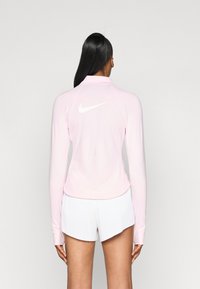 Bluză lungă cu mâneci, de culoare roz deschis, cu guler înalt și logo-ul alb Nike pe spate. Textură netedă și design ajustat, asortată cu pantaloni scurți albi.
