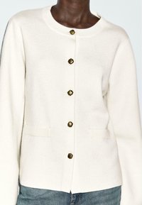 Cremefarbener Cardigan aus weichem Stoff, mit Rundhalsausschnitt, fünf goldfarbenen Zierknöpfen und zwei Taschen vorne.