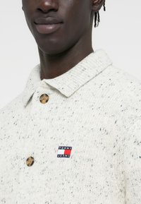 Ανοιχτό γκρι πουλόβερ με κολάρο, διακοσμημένο με ένα μεγάλο κουμπί και ένα μικρό λογότυπο "TOMMY JEANS" στο μπροστινό μέρος.