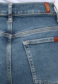 Οπίσθια όψη μπλε τζιν με θηλιές για ζώνη, λεπτομέρειες ραφών και πίσω τσέπη με μικρή καφέ ετικέτα.