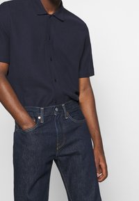 Man som bär en mörk marinblå kortärmad knapp-up skjorta instoppad i mörkblå denimjeans, med vänster hand i fickan mot en enkel bakgrund.