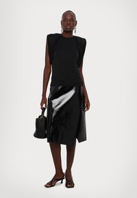 Schwarzes Kurzarmoberteil mit gerafftem Schulterdesign, kombiniert mit einem glänzenden schwarzen A-Linien-Rock. Die Accessoires umfassen eine schwarze Handtasche und Sandaletten.