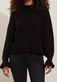 Pull noir en tricot avec un motif en diamant texturé, ourlet côtelé et poignets volants. Dispose d'un col montant et d'une coupe ample.