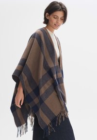 Wool blend poncho in brown with navy and beige check pattern, featuring a shawl collar and fringed edges for texture.