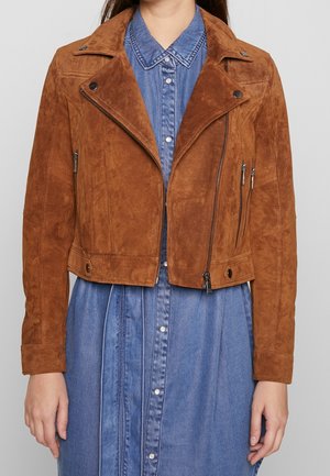 Femme portant une veste biker en daim marron par-dessus une robe en denim bleue boutonnée, debout devant un fond gris clair uni.