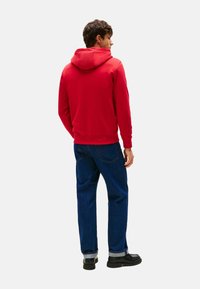 Hombre con sudadera roja, jeans azul oscuro y zapatos negros, de pie de espaldas contra un fondo claro y liso.
