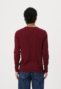 Bordeaux gebreide trui met lange mouwen, gestructureerde afwerking, geribbelde manchetten en zoom, gedragen over blauwe jeans, gezien vanuit de achterkant.