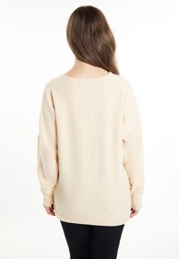 Beige, oversized pulóver, texturált, puha anyagból, széles ujjakkal és kerek hátú nyakkal. Fekete nadrággal párosítva.