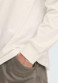 Camisa de algodón blanca de mangas largas con puños acanalados, combinada con pantalones verde oliva. Pulsera de cadena de plata visible en la muñeca.