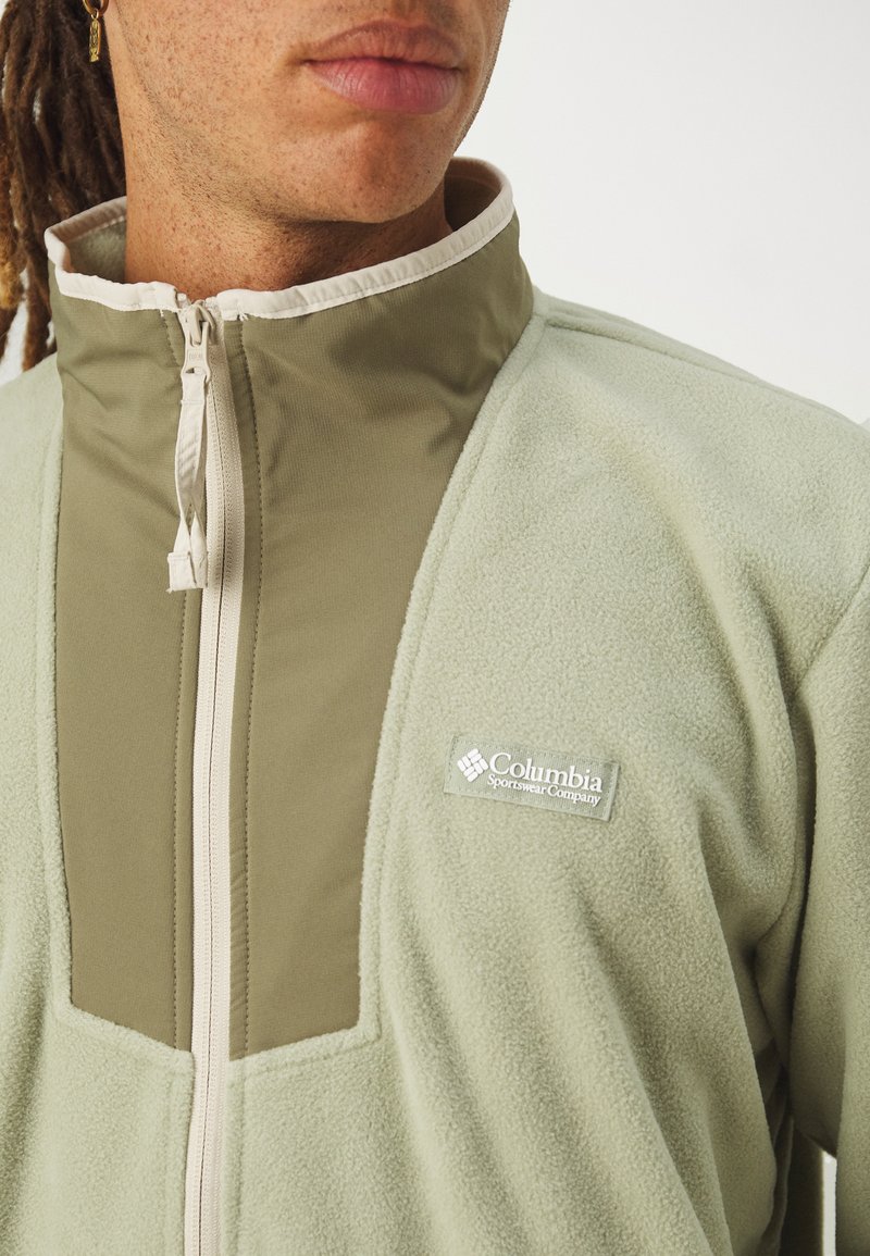 Lichtgroene fleecejack met een tan bovenpaneel, hoge kraag en een voorrits. Heeft een Columbia-logo op de linkerkant van de borst.