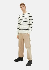Wit gestreepte trui met zwarte horizontale lijnen, gecombineerd met beige cargobroek met zakken en zwarte veterschoenen.