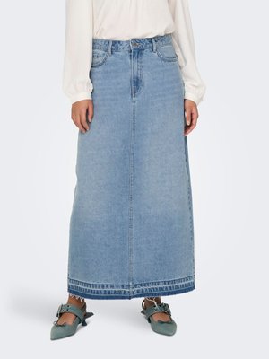 Femme porte une longue jupe en jean bleu clair avec un ourlet effiloché, un chemisier blanc et des chaussures à talons bleu sarcelle avec des boucles.