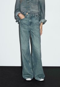 Person wearing loose-fitting faded blue jeans and a rolled-up denim shirt with one hand in pocket, standing against white wall.