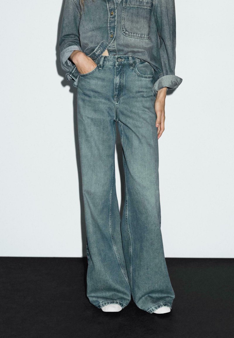 Person wearing loose-fitting faded blue jeans and a rolled-up denim shirt with one hand in pocket, standing against white wall.