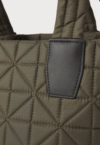Quilted olive green fabric tote bag with geometric pattern and black leather patch accent at the handle attachment. Textured surface detail visible.