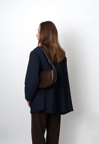 Borsa a tracolla con forma arrotondata, realizzata in pelle marrone scuro con linee verticali, abbinata a un blazer oversize blu navy e pantaloni marroni.