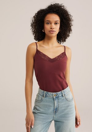 Žena s kučeravými vlasmi, nosiaca zlaté kruhové náušnice, bordový top s čipkovým lemom a svetlomodré rifle s vysokým pásom.