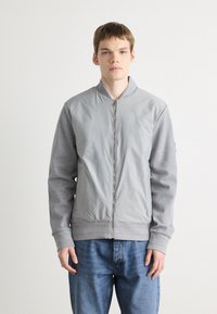 Jachetă bomber gri cu un front neted și mâneci texturate, cu manșete și tiv cu nervuri, completată de un sistem de închidere cu fermoar.