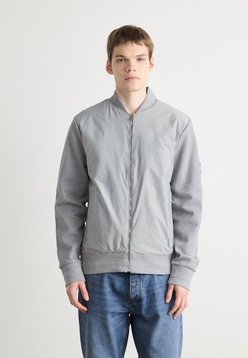 Jachetă bomber gri cu un front neted și mâneci texturate, cu manșete și tiv cu nervuri, completată de un sistem de închidere cu fermoar.