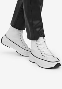 Sneaker alte bianche con suola in gomma spessa, caratterizzate da rifiniture nere, occhielli in metallo e lacci bianchi su un materiale in tela liscia.