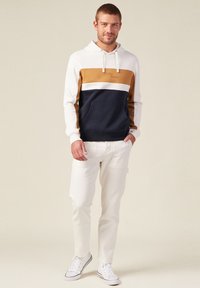 La felpa con cappuccio presenta un design a blocchi di colore con sezioni bianche, beige e blu navy; tasca a marsupio; abbinata a pantaloni cargo bianchi e sneakers.