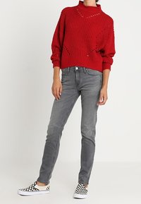 Pull rouge en tricot avec un col montant et des détails découpés, associé à un jean skinny gris et des chaussures slip-on à damier noir et blanc.