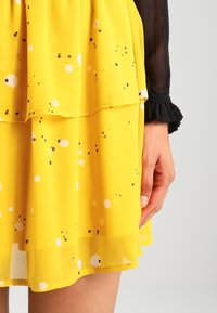 Abito giallo a strati con un design testurizzato. Presenta punti multicolori, sparsi di bianco, nero e blu su un tessuto fluente.