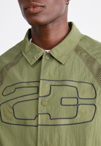 Ελαιόλευκο σακάκι με υφή υφάσματος, διαθέτοντας επενδύσεις από δίχτυ στους ώμους και μια εμφανή σχεδίαση λογότυπου με μαύρο περίγραμμα.