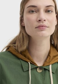 Veste verte avec une capuche marron, dotée d'une fermeture à boutons et de cordons de serrage. Le tissu semble lisse avec une texture légèrement brillante.