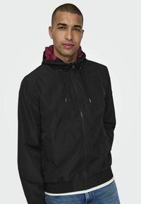 Zwarte zip-up jas met een rode gevoerde capuchon, elastische manchetten en tailleband. Lichtgewicht materiaal met trekkoorden voor een aanpasbare pasvorm.