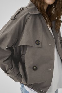 Frau trägt einen grauen doppelreihigen Trenchcoat über weißem Hemd und hellblauer Jeans, mit den Händen in den Manteltaschen.