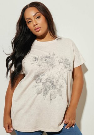 Kvinde med langt mørkt hår iført beige t-shirt med blomsterprint og korte ærmer samt blå jeans foran en ensfarvet lys baggrund.