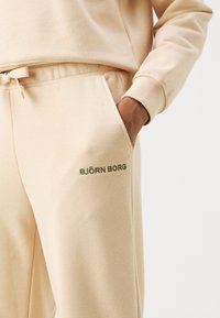 Beige joggingbroeken met een gestructureerde afwerking, voorzien van een trekkoord in de taille en een groen "BJÖRN BORG" logo op de zijzak.