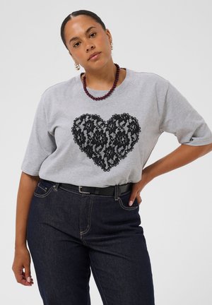 Kvinde iført en grå t-shirt med sort blondehjerte-design, mørke jeans med sort bælte, lilla perlehalskæde og creoløreringe.