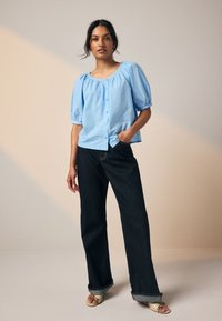 Hellblaue Bluse mit Puffärmeln und Knöpfen vorne, kombiniert mit dunkelblauen Jeans mit weitem Bein und beigen Sandalen mit offenen Zehen.