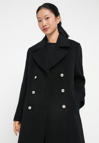 Manteau croisé en laine noire avec de grands revers, boutons argentés et poches latérales. Texture lisse avec une coupe décontractée.