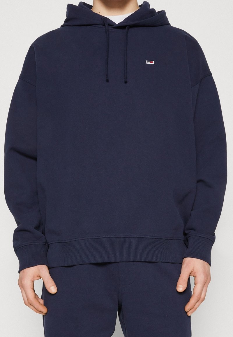 Marinblå hoodie tillverkad av mjukt tyg, med dragsko på huvan, ribbade cuffs och en liten logotyp på bröstet.