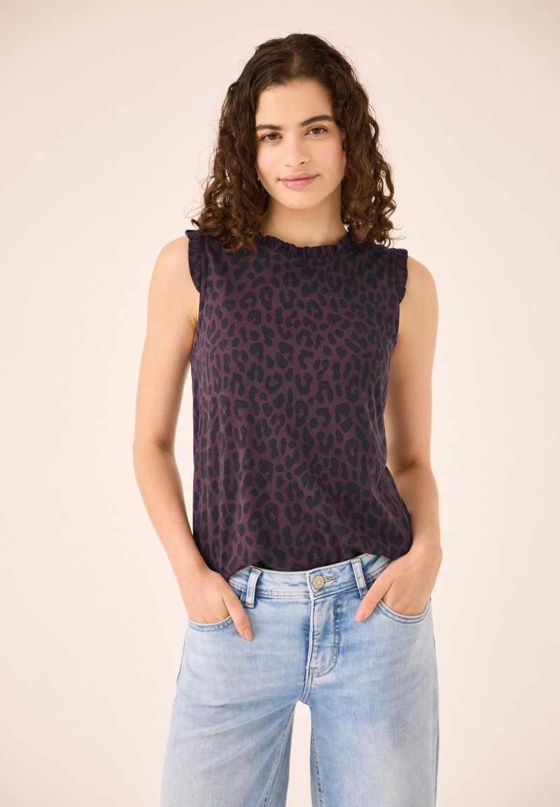 Jeune femme aux cheveux bouclés portant un top sans manches sombre à motif léopard et un jean bleu clair, mains dans les poches, sur un fond uni.