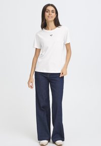 Fehér pamut póló kerek nyakkivágással és egy kis fekete szívmintával a mellrészen, széles lábú kék farmernadrággal párosítva.