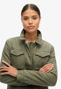 Veste militaire olive verte avec col montant, deux poches poitrine et accents de boutons, fabriquée en tissu solide.