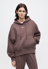 Νέος με κοντά σκούρα μαλλιά που φοράει μια υπερμέγεθη μωβ γιλέκο και αντίστοιχες παντόφλες, στέκεται σε έναν απλό λευκό φόντο.