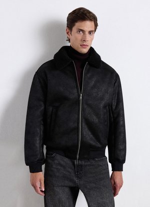 Junger Mann trägt eine schwarze Bomberjacke mit Lammfellkragen, dunklem Rollkragenpullover und ausgewaschenen schwarzen Jeans, steht an einer weißen Wand.