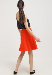 Haut sans manches noir associé à une jupe rouge évasée. La jupe est de longueur genou avec une texture lisse et un design simple. Des ballerines noires sont portées.